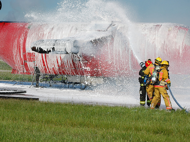 Firefighters in full protective gear spraying AFFF aqueous film-forming foam on an aircraft during a fire suppression training exercise — the source of PFAS contamination at military and airport sites