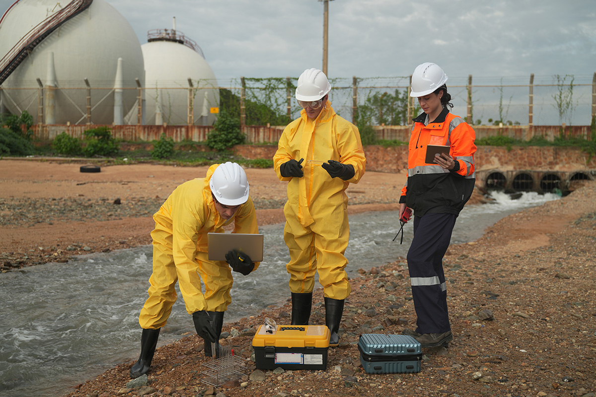 Environmental scientists in protective gear conducting water quality sampling near an industrial facility — field investigation at a PFAS-contaminated groundwater site