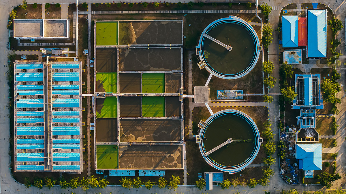 Aerial view of a municipal wastewater treatment plant with circular clarifier tanks and rectangular aeration basins â€” PFAS that enter through industrial and consumer sources concentrate in sewage sludge during the treatment process